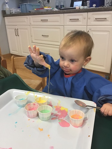 A toddler girl is using a spoon to scoop up goop.