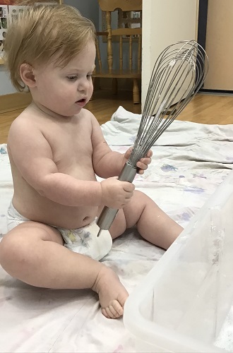 An infant is holding up a whisk, inspecting it closely.