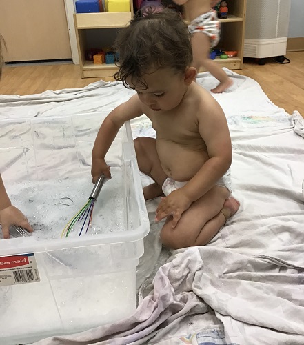 An infant is using a whisk in soapy water.