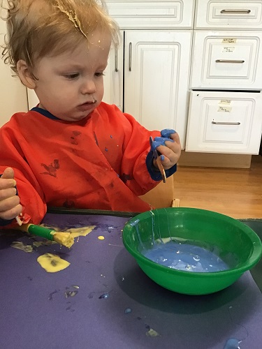 An infant has dipped their fingers into some blue paint and is inspecting them closely.