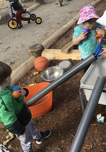 Some preschoolers are holding colourful balls as another preschooler pours water into a pipe to fill their container.