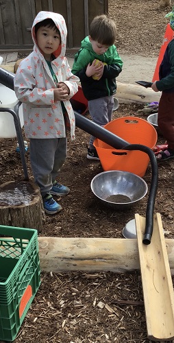 Preschoolers are standing infront of their containers and pipes they are using to fill them with water.