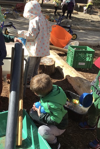 Some preschoolers are pouring water into pipes and collecting the water in bins.