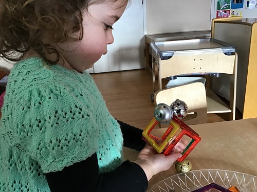 A preschooler is holding a magnetic tile block with metal jingle bells stuck to it.