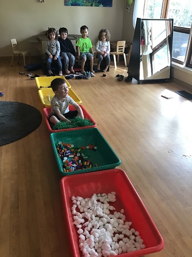 A preschooler is sitting in a sensory bin with loose parts inside as a group waits for their turn.