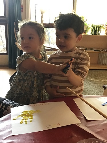 Two preschoolers each have different coloured paint on their hands and are pressing them together.