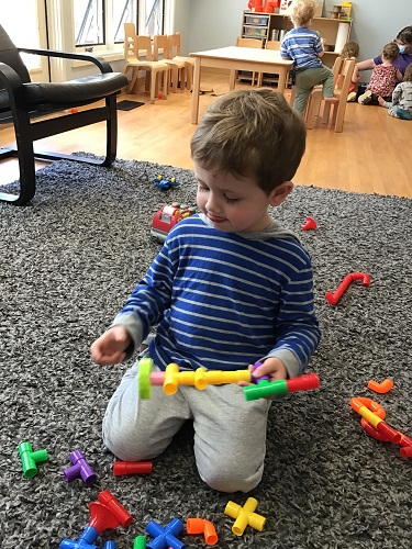 A toddler is putting tube blocks together as they sit on the carpet.