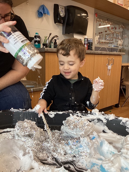 An educator adding glitter to the shaving cream while a child watches it with a smile