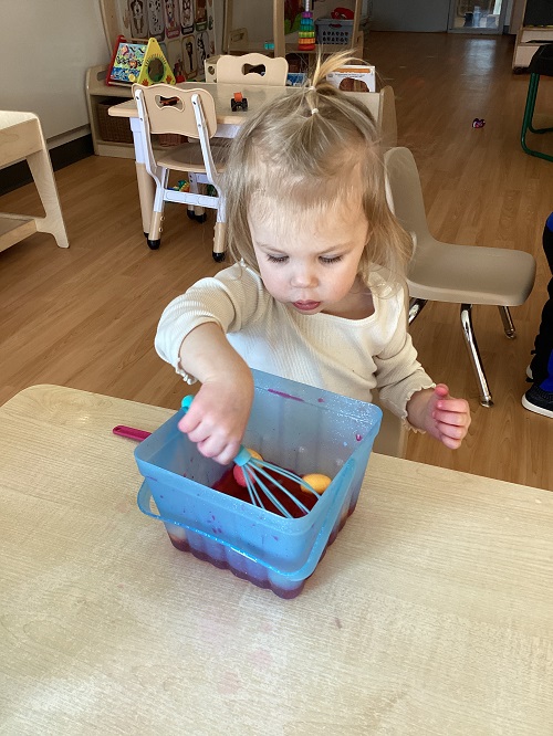 A toddler holding a whisk inside a container with coloured water and pretend eggs