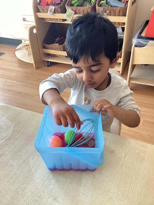 A toddler child holding a whisk inside a container with coloured water and plastic eggs