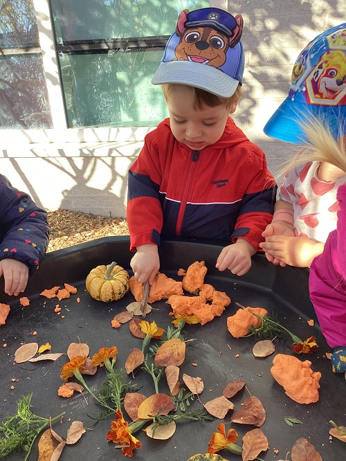 A toddler using a stick to cut orange play dough