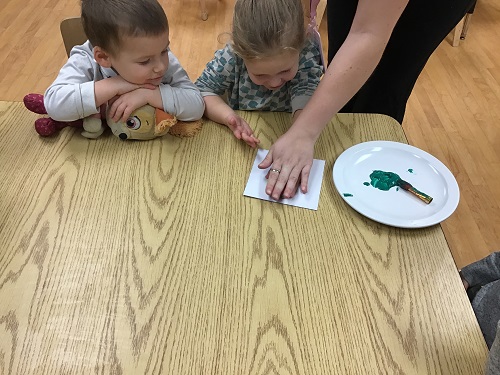 1 preschool child placing a painted handprint on paper and another preschool child observing