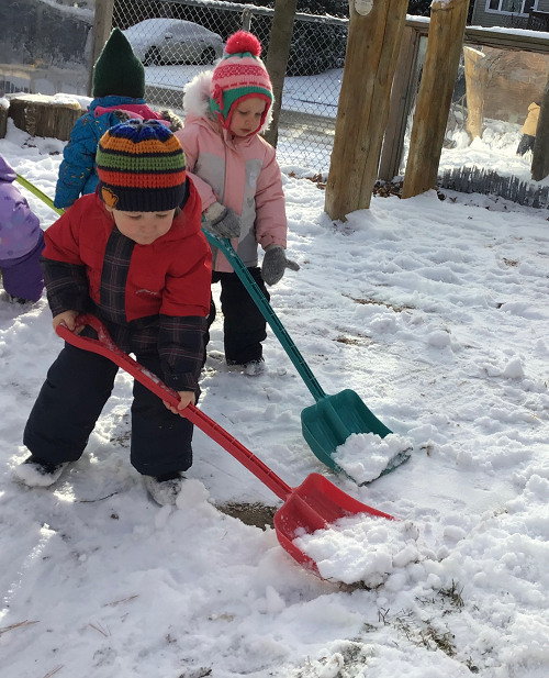 2 toddler children shoveling snow, one in the back observing